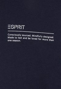 Námořnická modrá látka s bílou vyšívanou logem "ESPRIT", doplněná textem o udržitelnosti a designové filozofii.