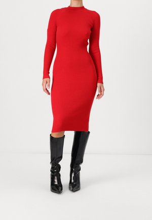 Vrouw in een nauwsluitende midi-jurk met lange mouwen in rood ribstof, gecombineerd met zwarte leren laarzen tot over de knie, staand tegen een eenvoudige achtergrond.