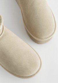 Beżowe zamszowe botki za kostkę z okrągłym noskiem i elastycznym panelem bocznym. Haftowane detale wzdłuż podeszwy i cholewki, gładka struktura na całej powierzchni.