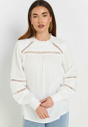 Blusa bianca con tessuto texturato, scollo arricciato e dettagli a taglio sulle spalle e sui polsini; presenta accenti plissettati lungo le maniche.