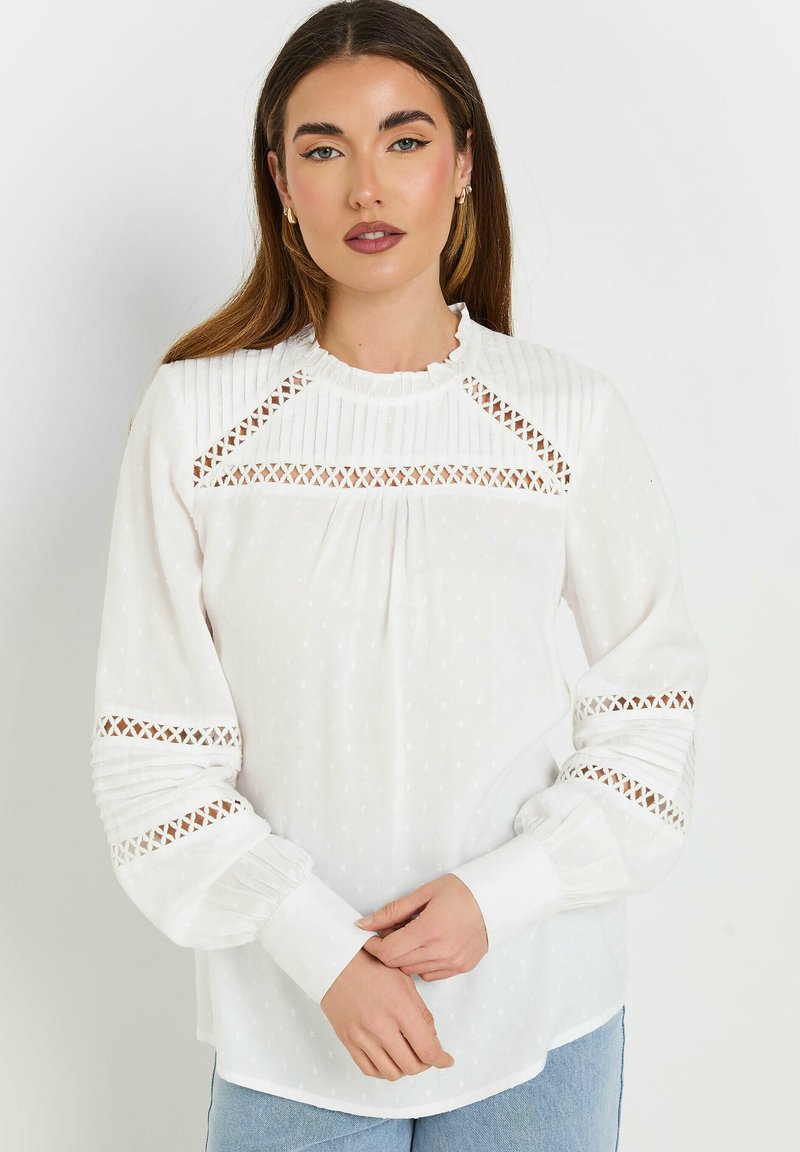Blusa bianca con tessuto texturato, scollo arricciato e dettagli a taglio sulle spalle e sui polsini; presenta accenti plissettati lungo le maniche.