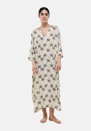 Femme debout pieds nus, portant une robe longue beige ample avec des imprimés noirs de palmiers et des manches trois-quarts.