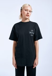 Černé bavlněné tričko s krátkými rukávy, s grafickým potiskem na levém prsu. Jednoduchý design s kulatým výstřihem a volným střihem.