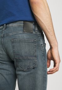 Man som bär blå jeans med synlig bakficka och svart lädermärke med varumärke på midjebandet, armen och handen avslappnade vid sidan.