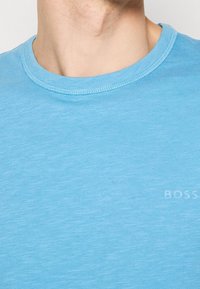 Gros plan d'une personne portant un T-shirt bleu clair à col rond avec un logo "BOSS" subtil sur la poitrine.