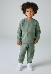 Conjunto de sudadera verde que incluye un sudadera de cuello redondo texturizada y joggers a juego. Detalle de logo notable en el pecho y los pantalones, con zapatillas blancas.