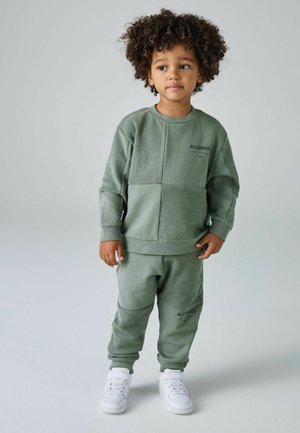 Zaļš sporta komplekts, kurā ietilpst teksturēts kreklu ar garām piedurknēm un saskaņotas sporta bikses. Ievērojams logo apzīmējums uz krūšu un biksēm, kopā ar baltām kedām.