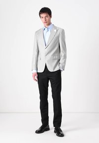 Lichtgrijze blazer, dichtgeknoopt met gepaspelde revers, gecombineerd met een blauw shirt en zwarte broek. Zwarte schoenen met een gladde textuur.