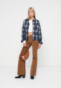 Niebieska i szara koszula w kratkę w oversize, biała krótka bluzka, brązowe spodnie w kant z grubego materiału, a także brązowa torebka o fakturowanej powierzchni z metalowymi elementami w kolorze srebrnym. Czarne kozaki na obcasie.