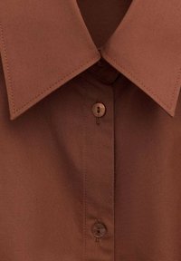 Chemise marron avec col pointu et deux boutons visibles sur la patte de boutonnage avant, montrant des coutures détaillées.