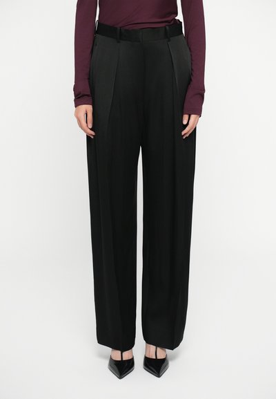 Pantalones negros de talle alto con plisados, confeccionados en una tela suave. Combinados con tacones negros de punta, creando una silueta a medida.