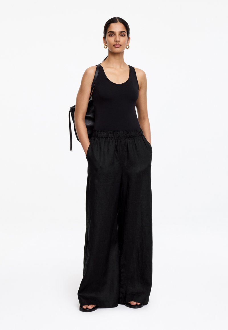 Femme debout, mains dans les poches, portant un débardeur noir sans manches, un pantalon noir à jambes larges, des boucles d'oreilles créoles et des sandales noires, sur fond blanc.