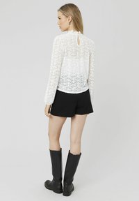 Haut en dentelle blanche avec des manches longues, détail en trou de serrure au dos, associé à un short noir et des bottes noires montantes sur un fond neutre.