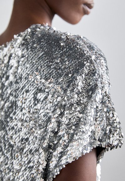 Silver sequin top with a textured surface, featuring a relaxed fit and short sleeves. The sequins reflect light, creating a shimmering effect.