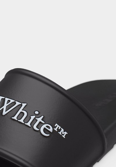 Μαύρο σανδάλι με σαγιονάρα με ανάγλυφο λευκό κείμενο «White™» στο λουράκι, σε κοντινή λήψη πάνω σε απλό φόντο.