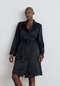 Svart satin morgonrock med långa ärmar, en sjalkrage och ett matchande bälte. Har en knälång nederdel och en mjuk, blank yta.