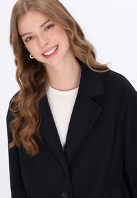 Giovane donna con lunghi capelli mossi di color castano chiaro, vestita con una maglietta bianca e un cappotto oversize nero, che sorride contro uno sfondo bianco uniforme.