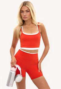 Jeune femme aux longs cheveux blonds portant une tenue de sport rouge et blanche, tenant une bouteille d'eau argentée et un casque blanc.