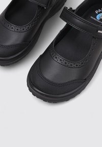 Zapatos negros estilo Mary Jane con correa ajustable y puntera texturizada sobre un fondo gris claro.