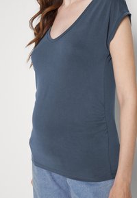 Camiseta azul de decote em V, feita de um tecido suave, com mangas curtas enroladas e detalhe de franzido lateral, apresentando uma textura lisa e um corte ajustado.