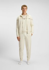 Lichtbeige ritsjack voor sport met bijpassende broek. De stof lijkt glad; het jasje heeft contrasterende strepen en het "BOSS"-logo op de borst.