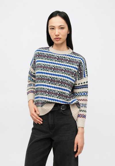 Woman with straight dark hair wearing a multicolored patterned knit sweater tucked into high-waisted black pants with a black belt.