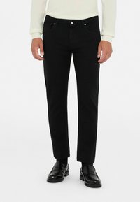 Mustad teksased, slim-fit lõikega, viie taskuga ja nööpidega kinni pandud. Kangas on siledast tekstuurist, kombineeritud mustade nahast loaferitega.