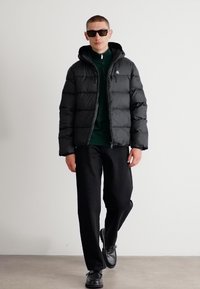 Zwarte pufferjas met capuchon, voorzien van een ritssluiting aan de voorkant en een doorgestikt ontwerp. Draag het over een groene ritsjack en combineer het met zwarte broek en schoenen.