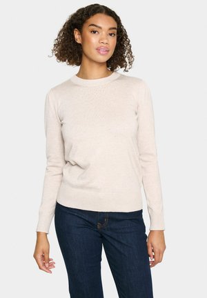 Pull en tricot beige clair avec un col rond, des poignets et un ourlet côtelés, présentant un motif subtil sur le devant. Porté avec un jean bleu foncé.