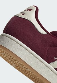 Maroon suede sneaker with a white leather heel tab, featuring textured rubber outsole and signature logo on the heel.