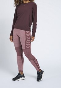 Długi rękaw bordowej aktywnej bluzki o gładkiej fakturze. Dopasowane legginsy z teksturą mają wzory w kształcie chevronów w odcieniach różu, zestawione z czarnymi trampkami.