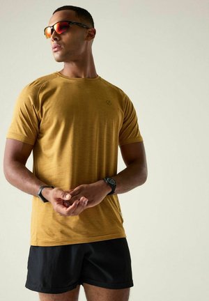 Mann trägt bernsteinfarbene Sportsonnenbrille, senffarbenes T-Shirt, schwarze Shorts und zwei Armbanduhren, steht vor einem schlichten hellen Hintergrund.