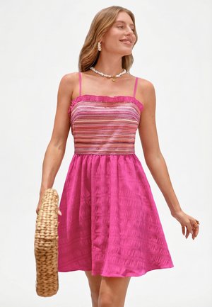Femme souriante portant une robe rose sans manches avec un corsage à motifs, tenant un sac panier tissé, debout devant un fond blanc uni.