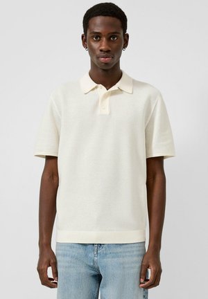 Junger Mann mit kurzen lockigen Haaren, der ein strukturiertes, elfenbeinfarbenes Poloshirt und hellblaue Jeans trägt, vor einem einfarbigen Hintergrund stehend.