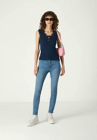 Γυναίκα με σγουρά μαλλιά, φορώντας μαύρα γυαλιά ηλίου, ναυτική αμάνικη μπλούζα, ανοιχτό μπλε τζιν, λευκά sneakers και κρατώντας μια ροζ τσάντα ώμου.