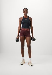 Black athletic tank top with purple accents, maroon shorts featuring "NIKE" logo, holding black dumbbells, wearing beige sneakers and white socks.