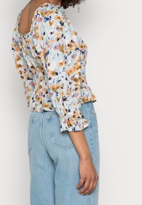 Blouse fleurie en tissu bleu clair avec manches bouffantes, taille smockée et dos carré. Associée à un jean en denim clair.
