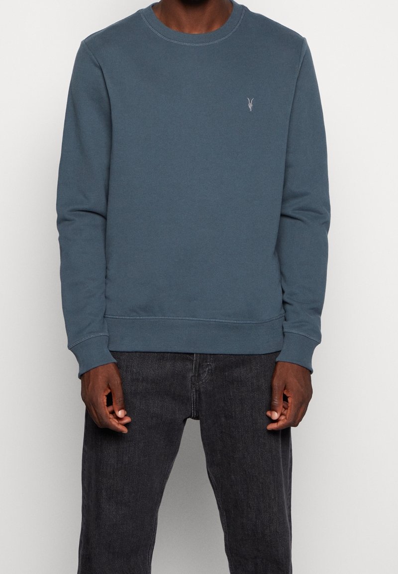 Homme portant un sweat-shirt bleu à manches longues avec un petit logo blanc sur la poitrine et un jean noir, debout devant un fond blanc uni.