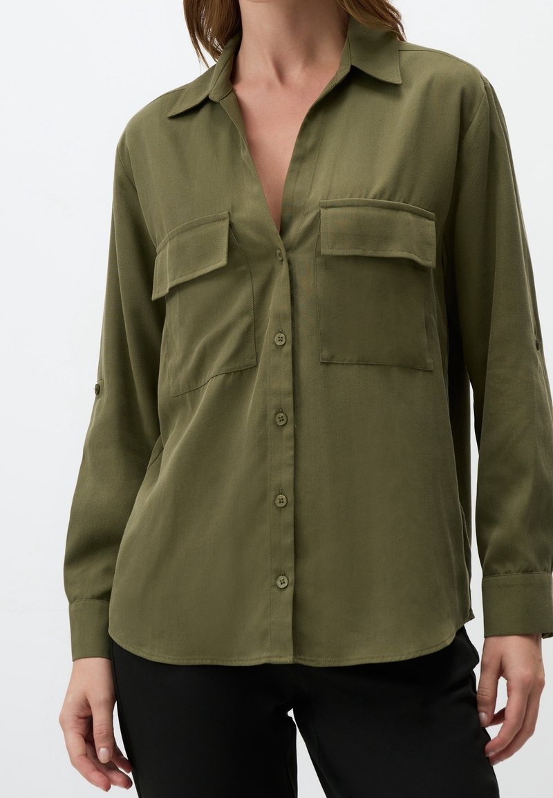Femme portant une chemise boutonnée vert olive avec deux poches poitrine et manches longues, associée à un pantalon noir, debout devant un fond blanc.