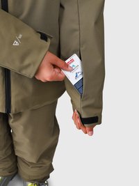 Persona che indossa una giacca e pantaloni da sci verde oliva mentre inserisce un biglietto per la risalita nella tasca interna della giacca, dotata di chiusura in velcro.