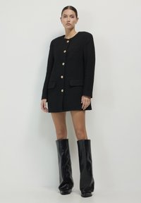 Abito blazer in bouclé nero con bottoni dorati, dotato di scollatura rotonda e tasche frontali. Stivali neri in pelle al ginocchio completano il look.
