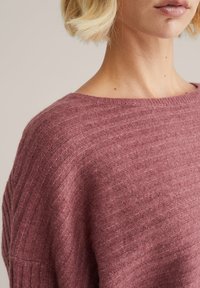 Mauve kötött pulóver bordázott textúrával, széles nyakkal és leengedett vállakkal, amely egy puha, könnyű anyagot mutat be.