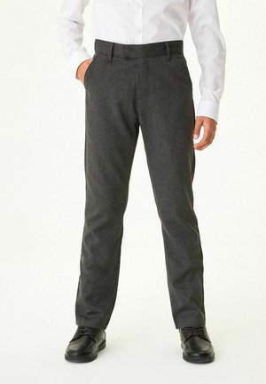 Homme portant un pantalon droit gris foncé, des chaussures noires et une chemise blanche à manches longues, debout avec une main dans la poche.