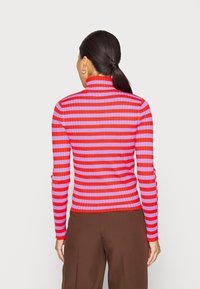 Kvinna med mörkt hår i en låg hästsvans, iklädd en röd och lila randig turtlenecktröja och bruna byxor, vänd bort.