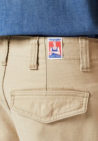 Pantaloni Wrangler beige con etichetta Casey Jones, caratterizzati da una tasca posteriore cucita e una superficie liscia. Camicia blu parzialmente visibile sopra.