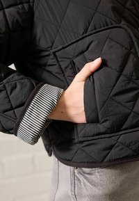 Prošívaná černá bunda s boční kapsou a pruhovaným manžetovým lemem. Bunda má hladkou texturu a přiléhavý střih.