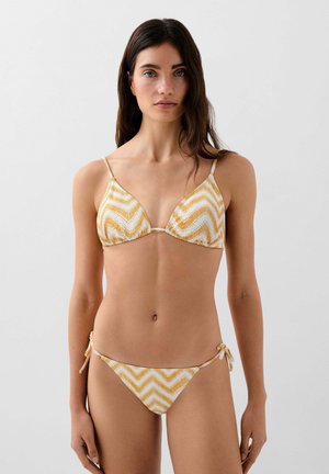 Mujer con cabello largo y oscuro que lleva un bikini de hilo con patrón de chevrón amarillo y blanco, mirando hacia adelante sobre un fondo liso.