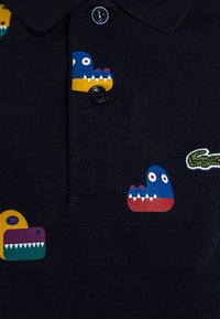Mörkblå polo-shirt med färgglada monstermotiv i blått, gult och rött, med svarta knappar och en grön broderad logotyp.