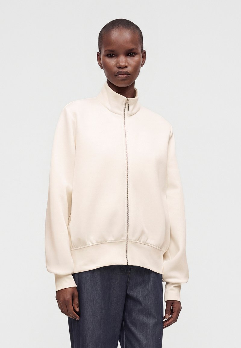 Personne portant une veste crème zippée avec un col haut et un pantalon foncé ample, debout devant un fond clair uni.