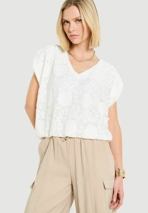 Blondynka w białej teksturowanej bluzce z krótkim rękawem, beżowych spodniach z wysokim stanem, złotym naszyjniku z łańcuszka i złotej bransoletce.